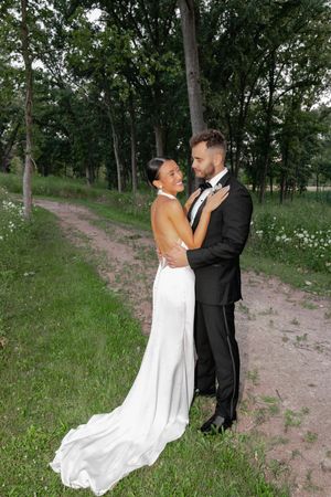 Bride in white gown and groom in black tuxedo embrace on a lush green path surrounded by trees.