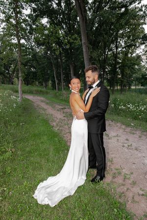 Bride in white gown and groom in black tuxedo embrace on a scenic garden path surrounded by lush green trees.
