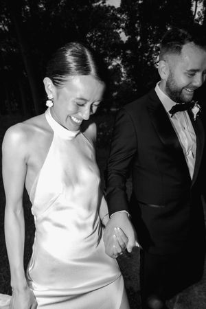 Black and white photo of a smiling bride in a halter gown holding hands with a tuxedo-clad groom at a wedding.