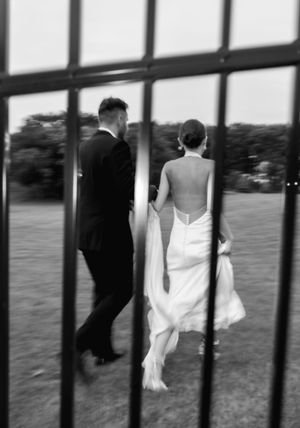 Black and white photo of bride and groom walking hand in hand through iron gates on wedding day.