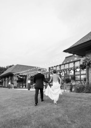 Black and white photo of a bride and groom holding hands running toward a venue with string lights at dusk.