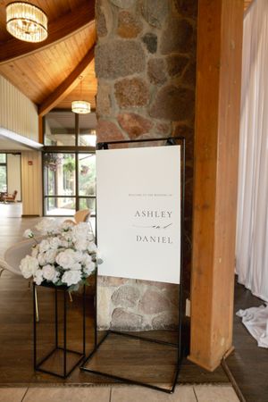 Elegant wedding welcome sign for Ashley and Daniel displayed on a black stand beside white floral arrangements in a rustic venue.