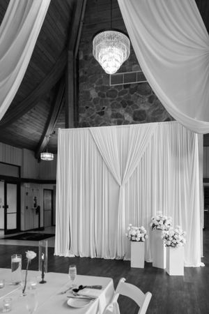 Elegant black and white wedding reception with white drape backdrop, floral arrangements, chandelier, and decorated tables.