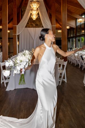 Bride in white halter gown holding white orchid bouquet, twirling in elegant wood-beamed wedding reception venue.