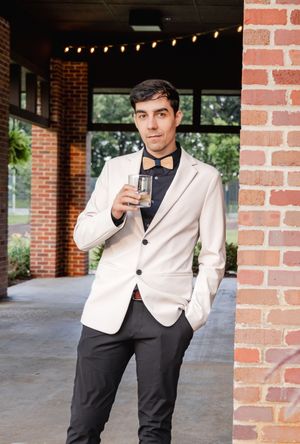 Young man in white blazer and bow tie holding a drink, posing by a brick column at an elegant outdoor venue.