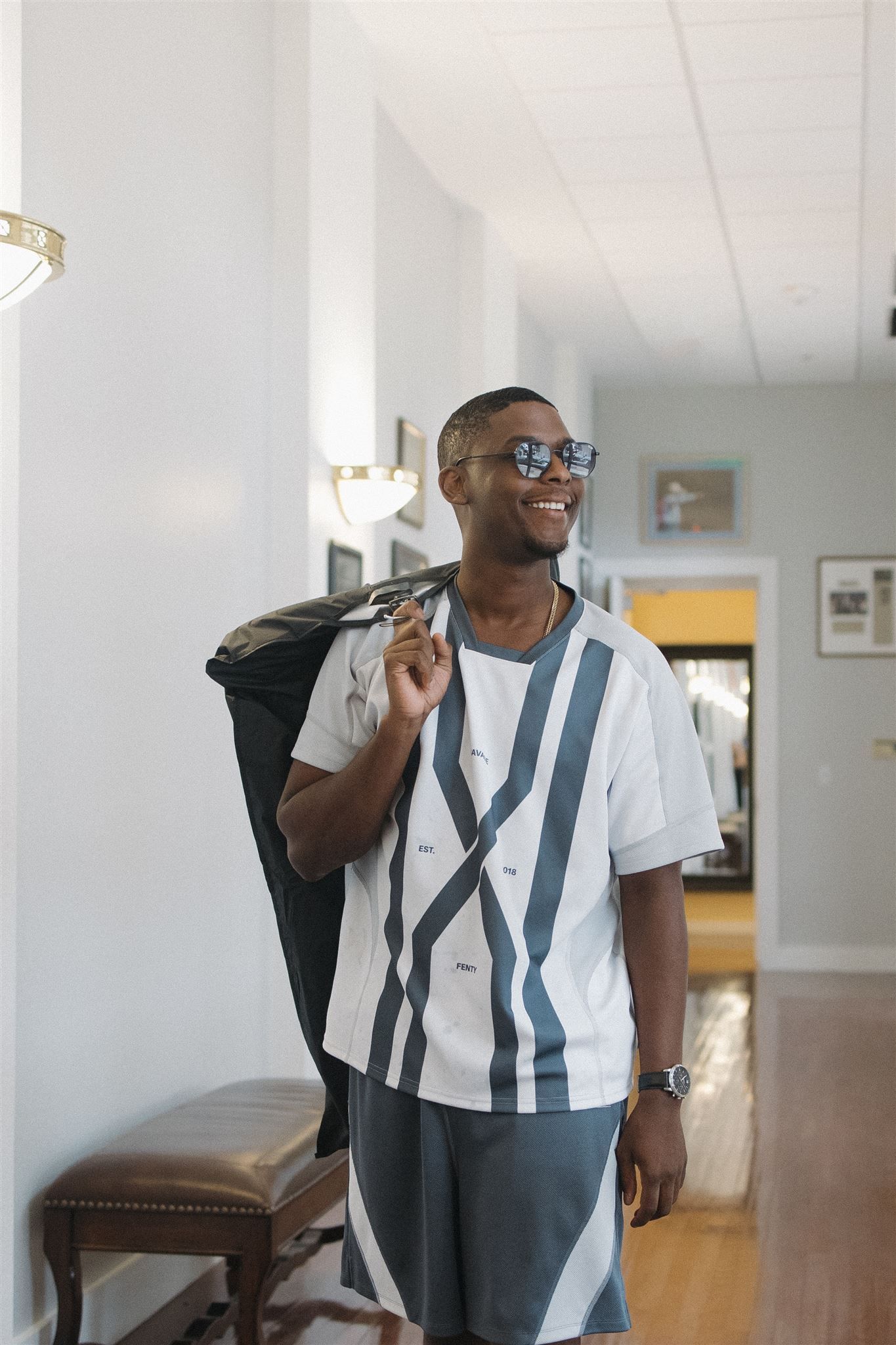 Smiling person in a white striped shirt and glasses holding a jacket while standing in a bright hallway interior.