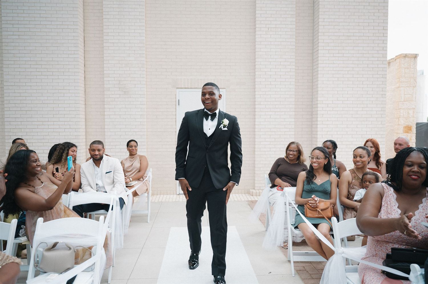 A groom walks down the aisle at a wedding ceremony while guests look on smiling in an elegant white venue.