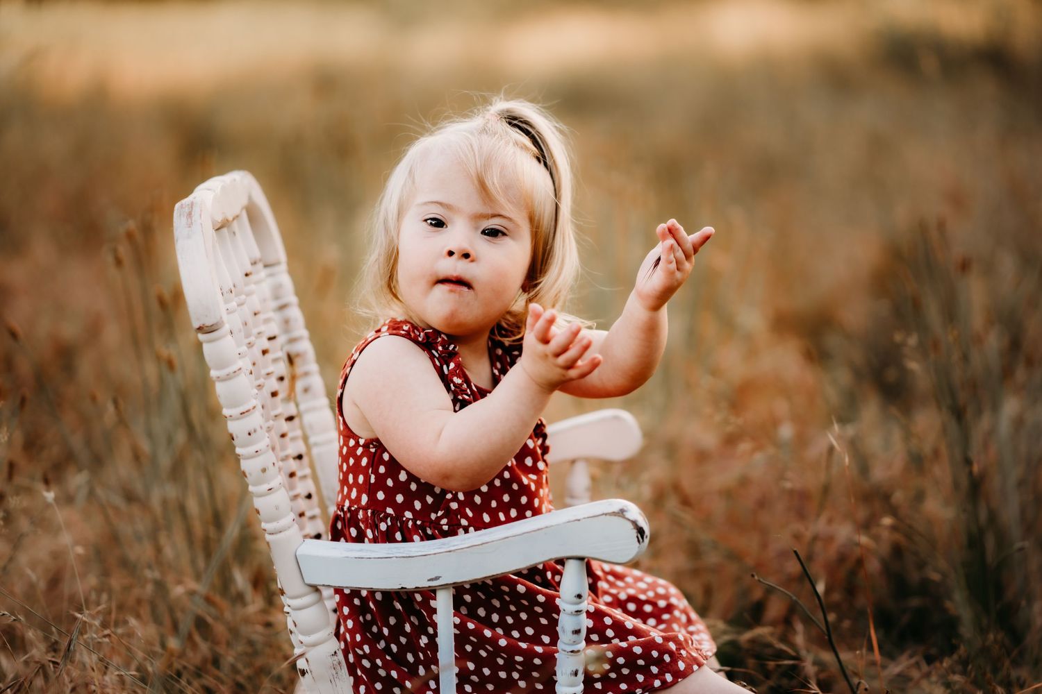 A small child in a red polka dot dress sits in a white antique chair amid golden autumn grass.