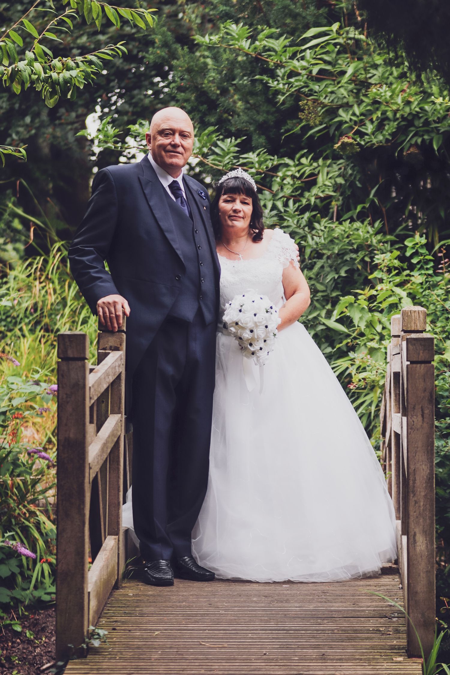 A couple poses on a wooden bridge surrounded by lush garden scenery on their wedding day.