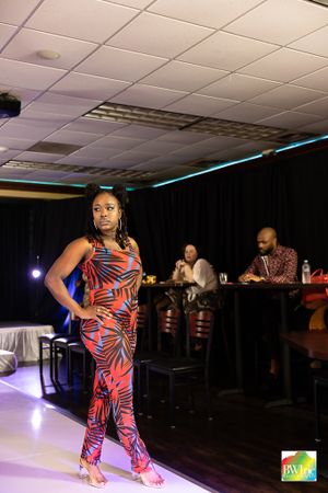 Runway model wearing red and black tropical print jumpsuit walks catwalk with audience in background.