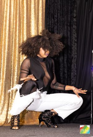 Performer in black mesh top and white pants strikes dynamic pose against gold curtain backdrop.
