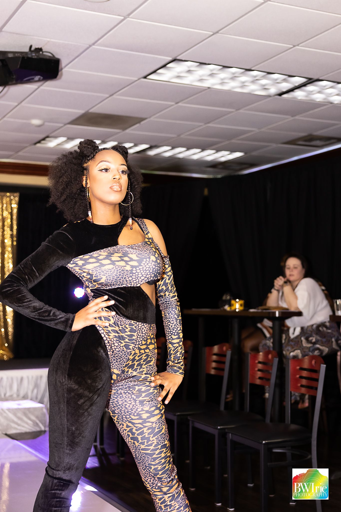 Confident model strikes pose in geometric patterned jumpsuit on illuminated runway with bar seating visible.