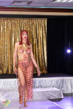 Red crystal-embellished costume with high slit shown on runway against gold curtain backdrop.