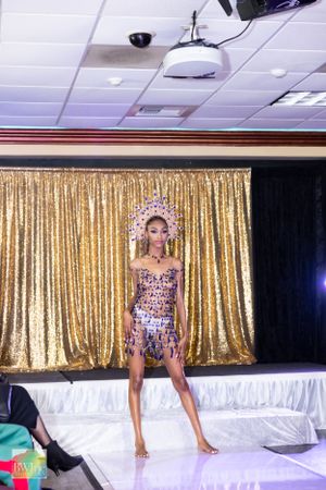 Crystal-adorned costume with crown headpiece sparkles against gold curtain backdrop on fashion runway.