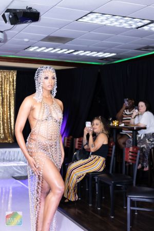 Model in sheer crystal bodysuit poses on runway with gold sequin backdrop and dramatic lighting.