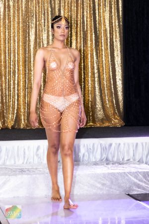 Fashion model in sheer peach crystal outfit poses on white runway with gold backdrop.