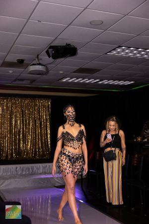 Fashion model walks runway in dark sequined outfit against gold curtain backdrop during nighttime fashion show.