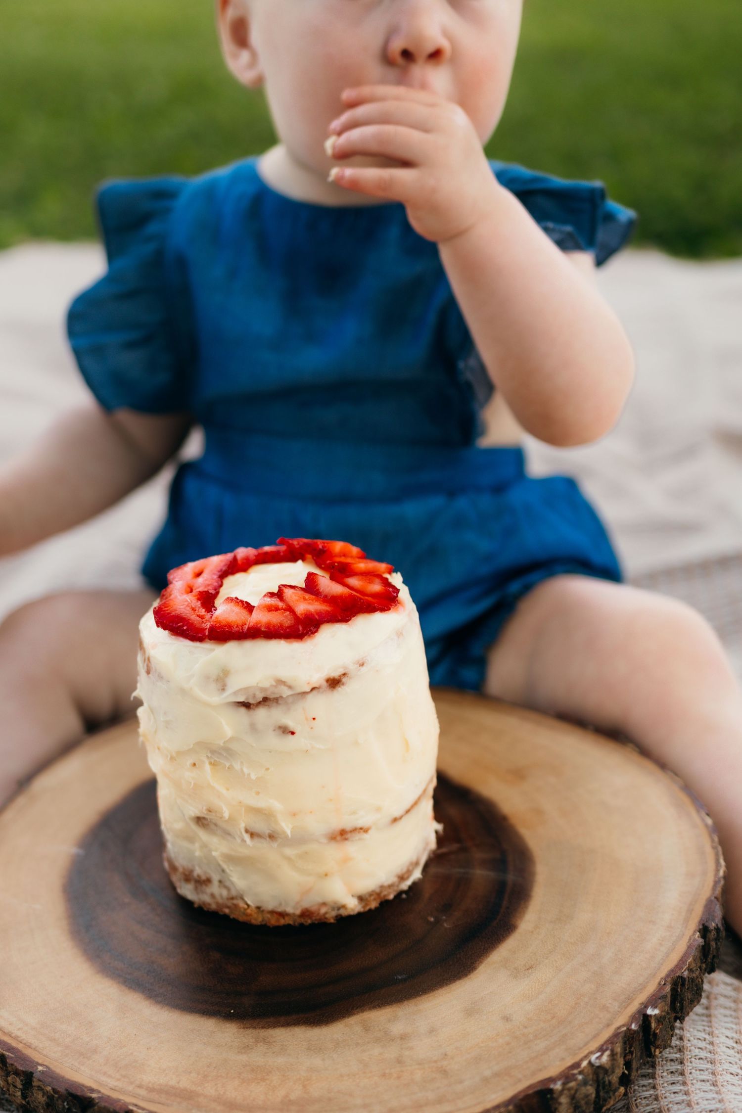A cake in the foreground with a young baby licking icing off of her hand in the background.