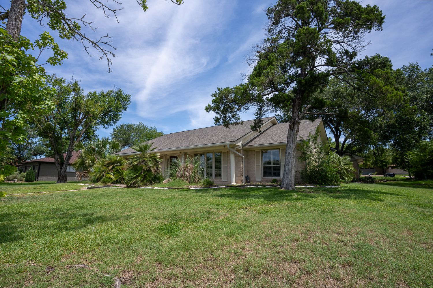 Single-story ranch house with white exterior and large front lawn surrounded by mature oak trees on a sunny day.