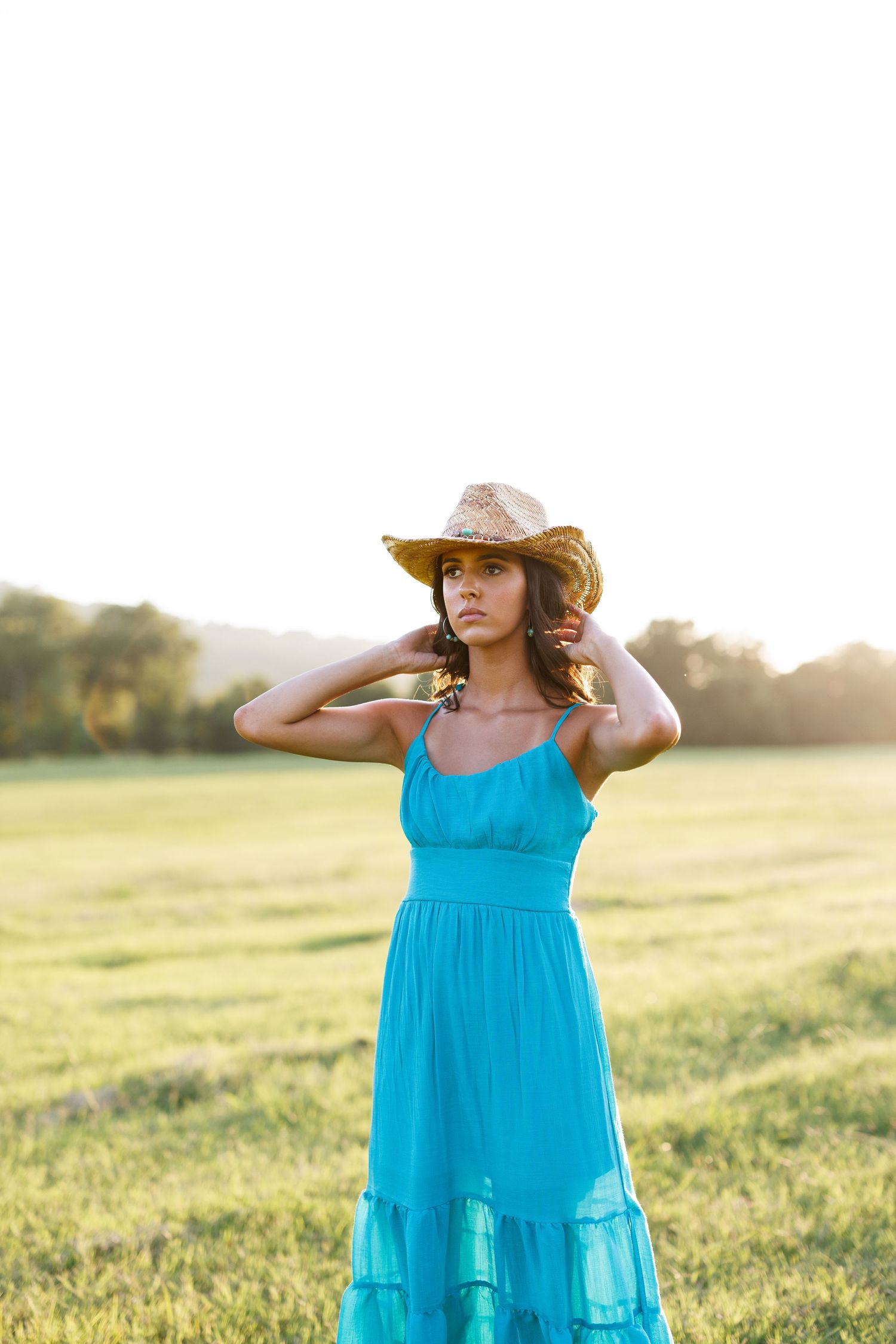 Woman in turquoise sundress and cowboy hat poses gracefully in sunlit meadow at golden hour.