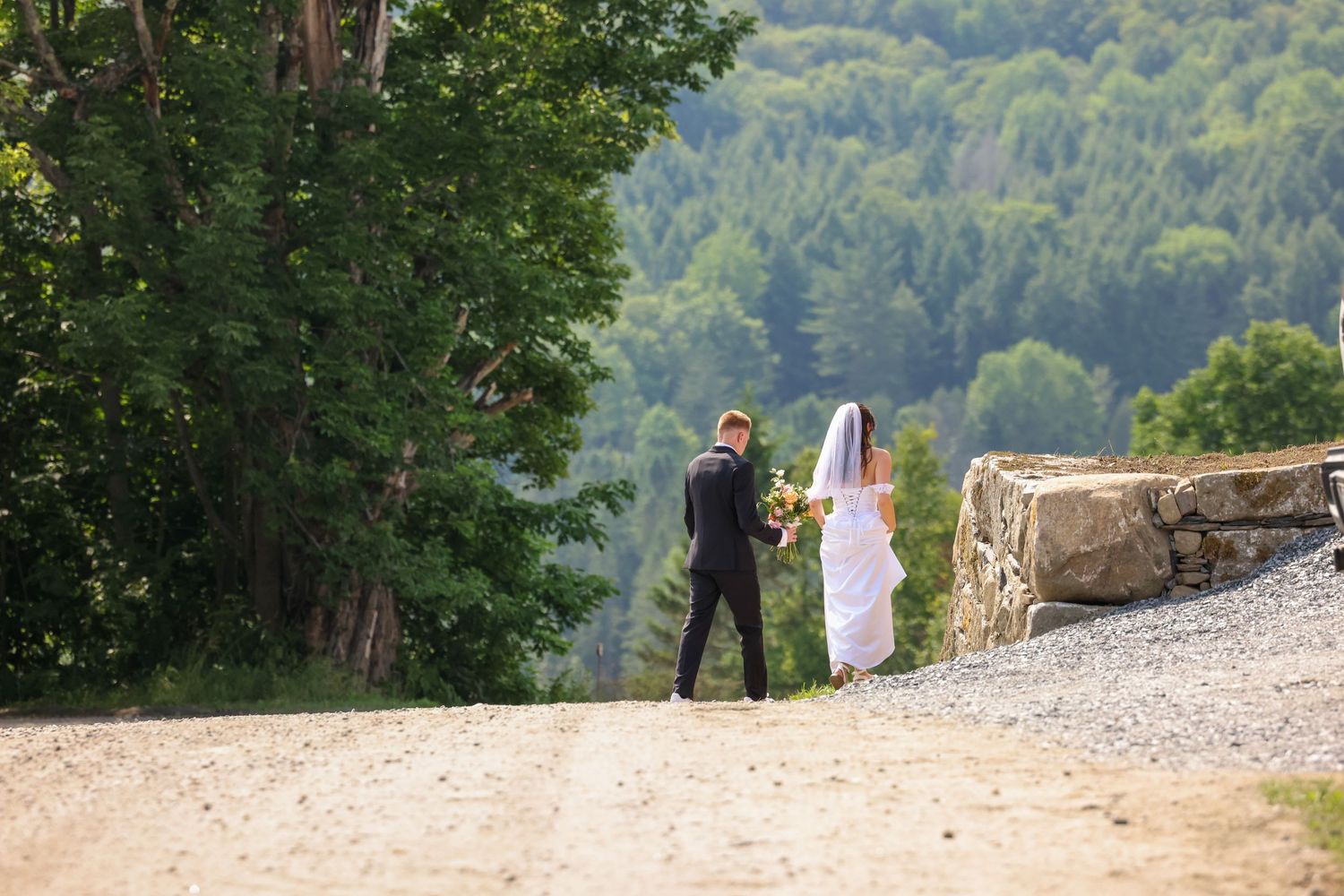 Bride and groom walking hand in hand on a gravel path surrounded by lush green forest.