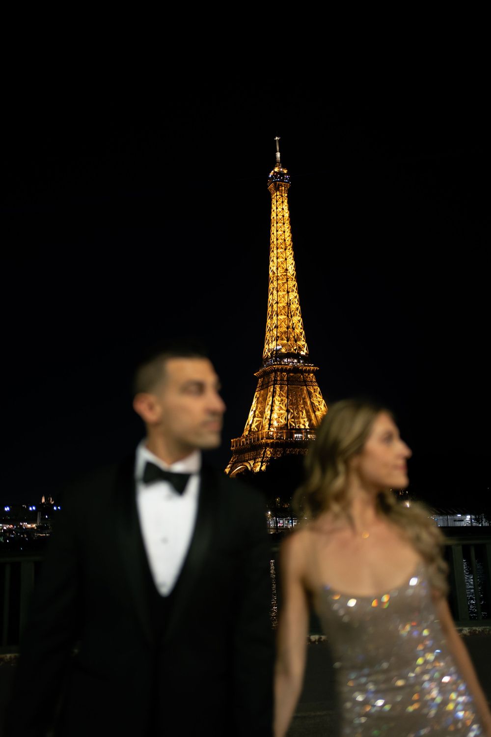 Elegant couple in formal attire with the illuminated Eiffel Tower glowing gold against the night sky in Paris.