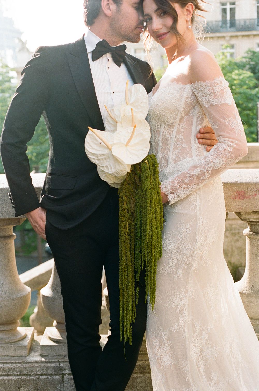 Bride in lace off-shoulder gown holds white anthurium bouquet beside groom in black tuxedo on stone balcony.