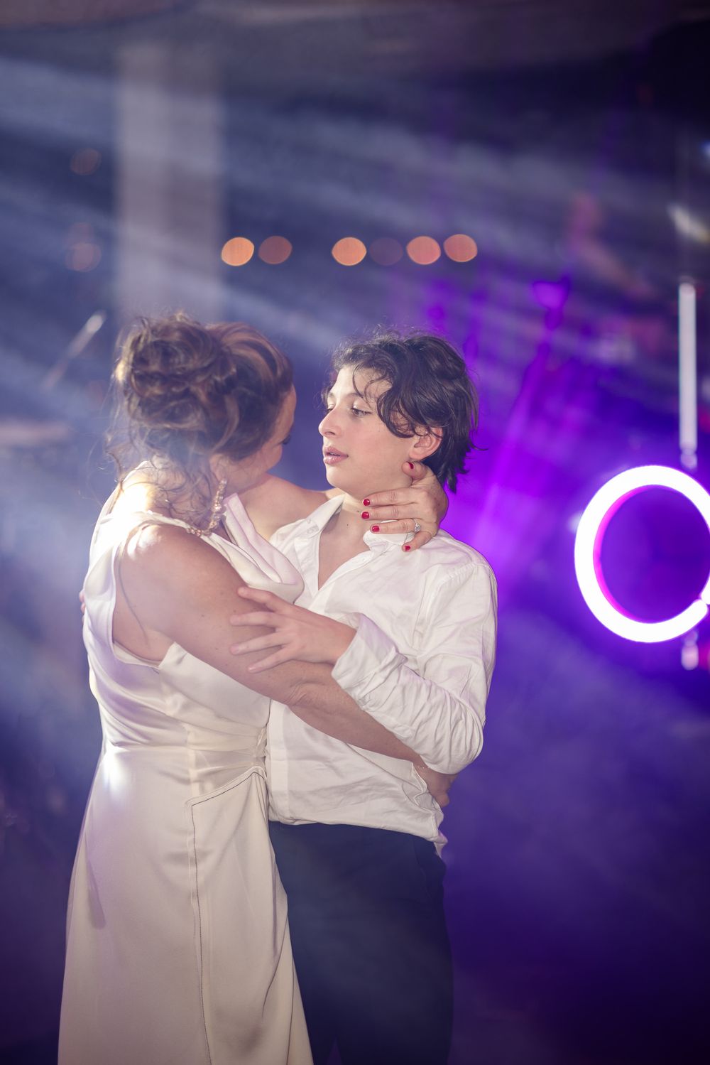 A couple shares their first dance at a wedding reception with purple lighting and bokeh effects in the background.