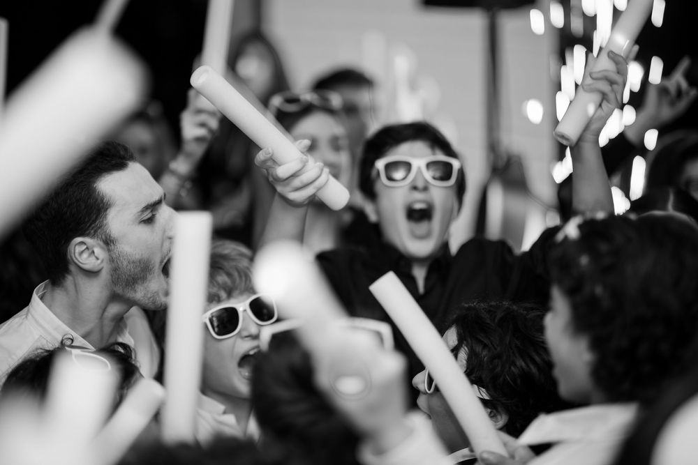 Energetic crowd of concert-goers dance and cheer with raised hands at a live music performance.