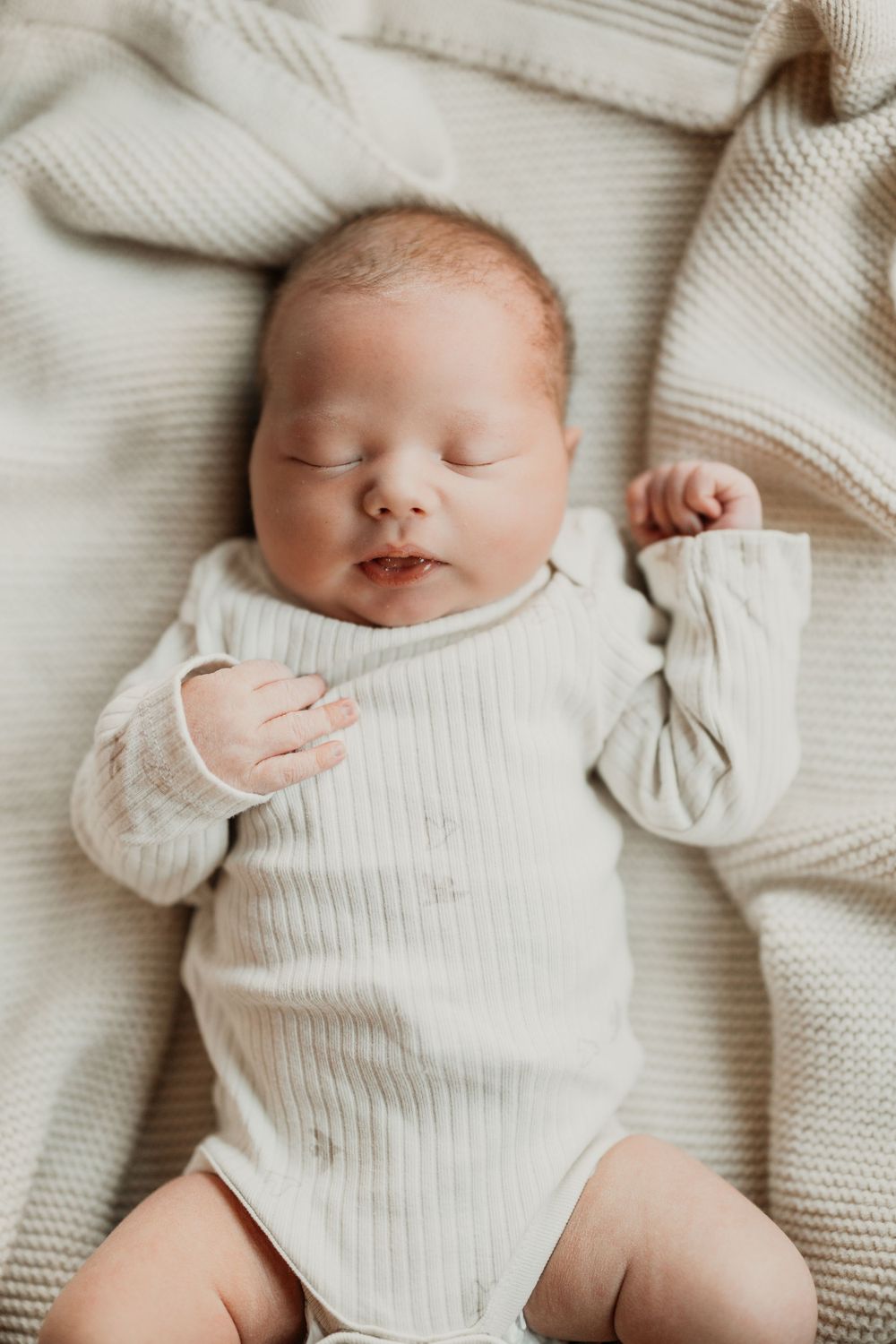 Sleeping baby in white outfit on cozy knit blanket with gentle natural lighting.