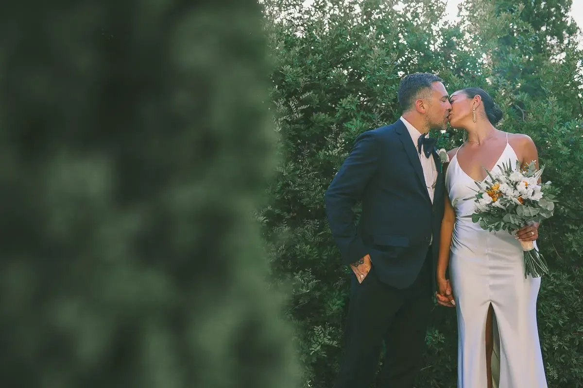 Bride and groom share a romantic kiss outdoors, surrounded by lush greenery, bride holding a bouquet.