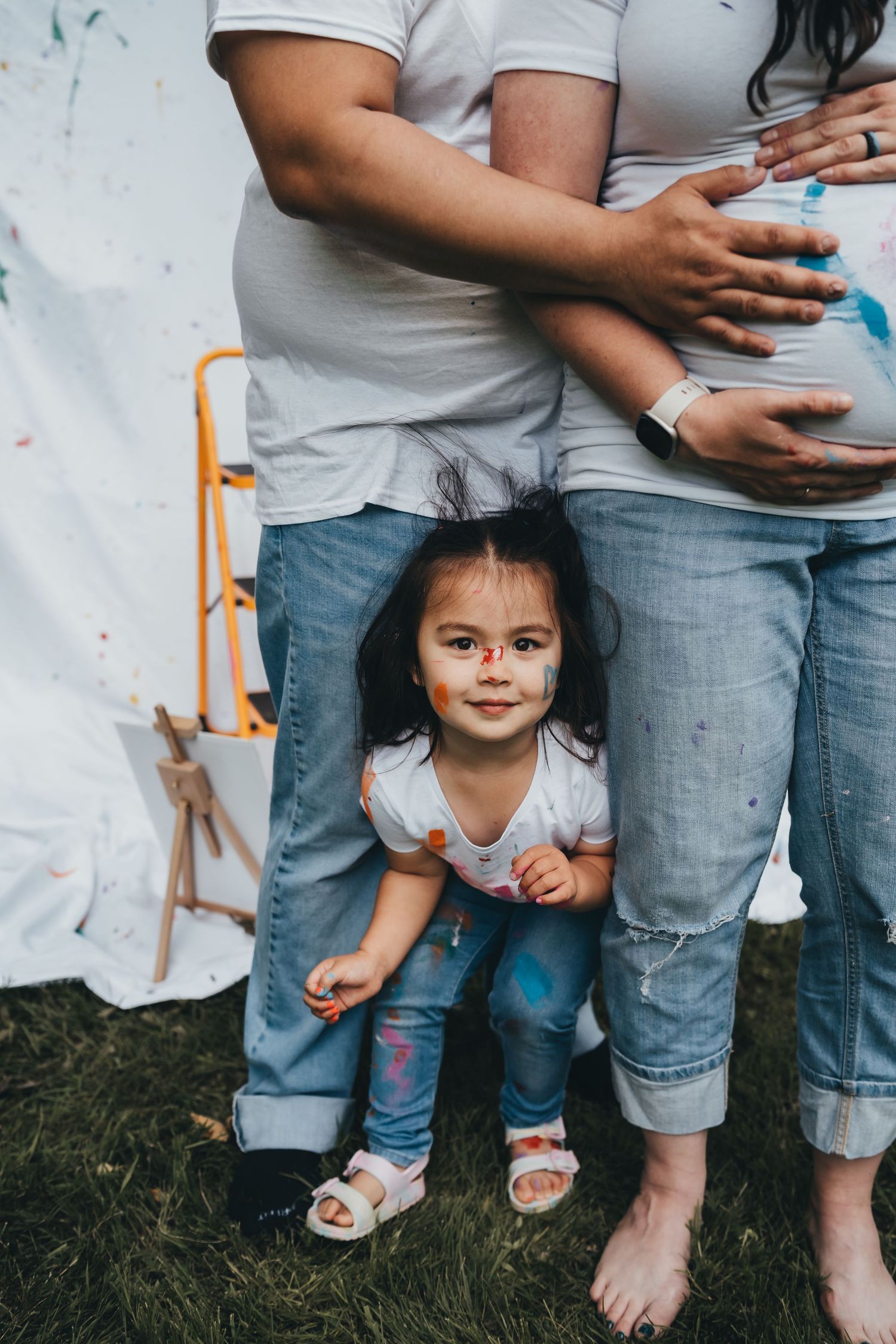 Young child peeks out playfully between parents' legs during outdoor photo session with white backdrop.