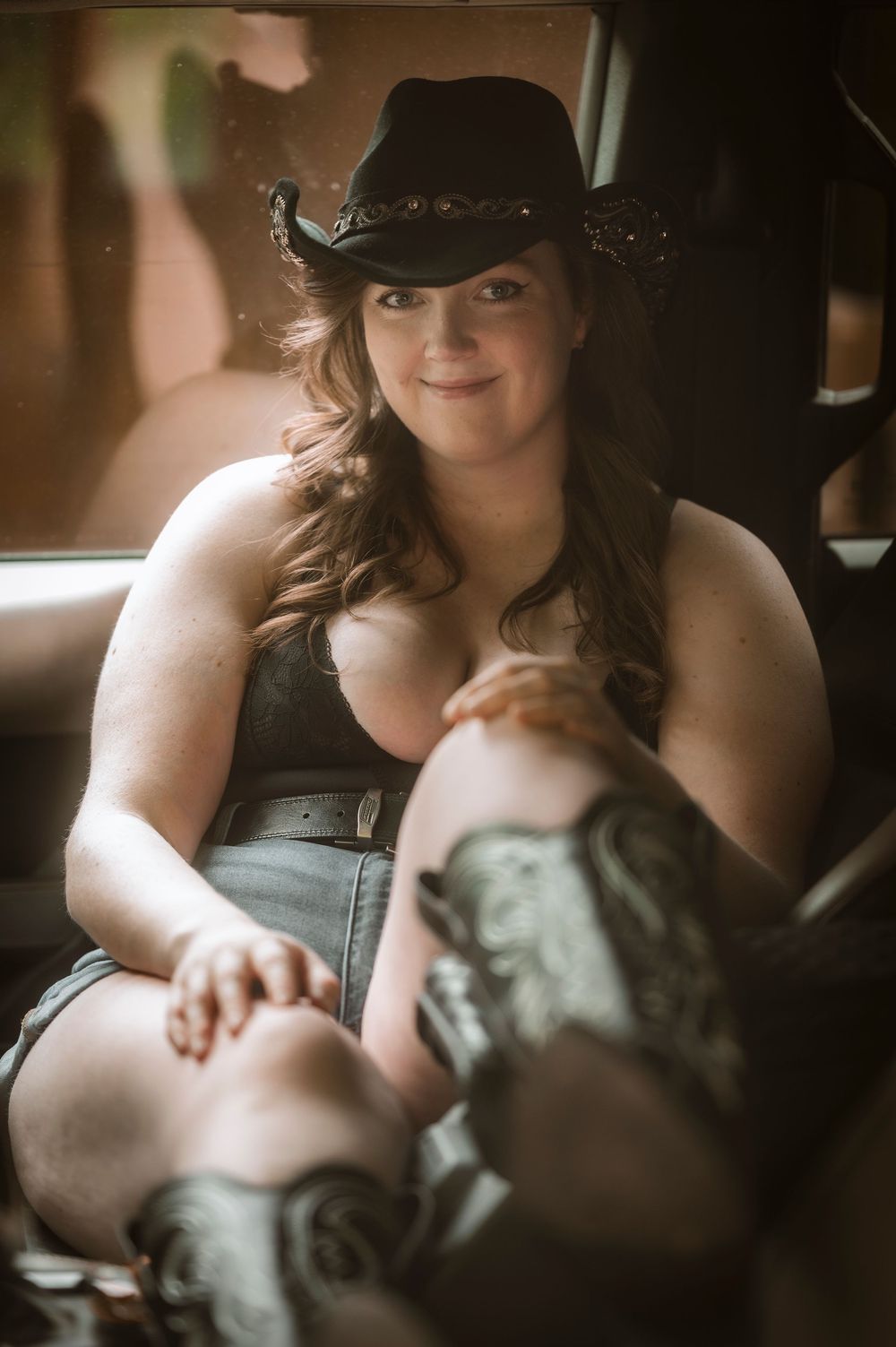 A smiling woman in a black cowboy hat, denim shorts, and cowboy boots sits relaxed inside a vehicle.