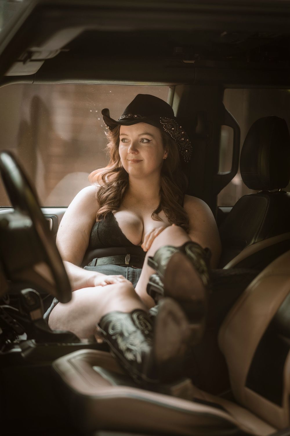 Woman in black cowboy hat and lingerie sitting in a vehicle, smiling with warm lighting.