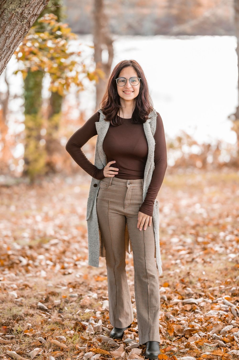 A person wearing brown sweater and gray dress pants poses in autumn leaves during a fall photoshoot outdoors.