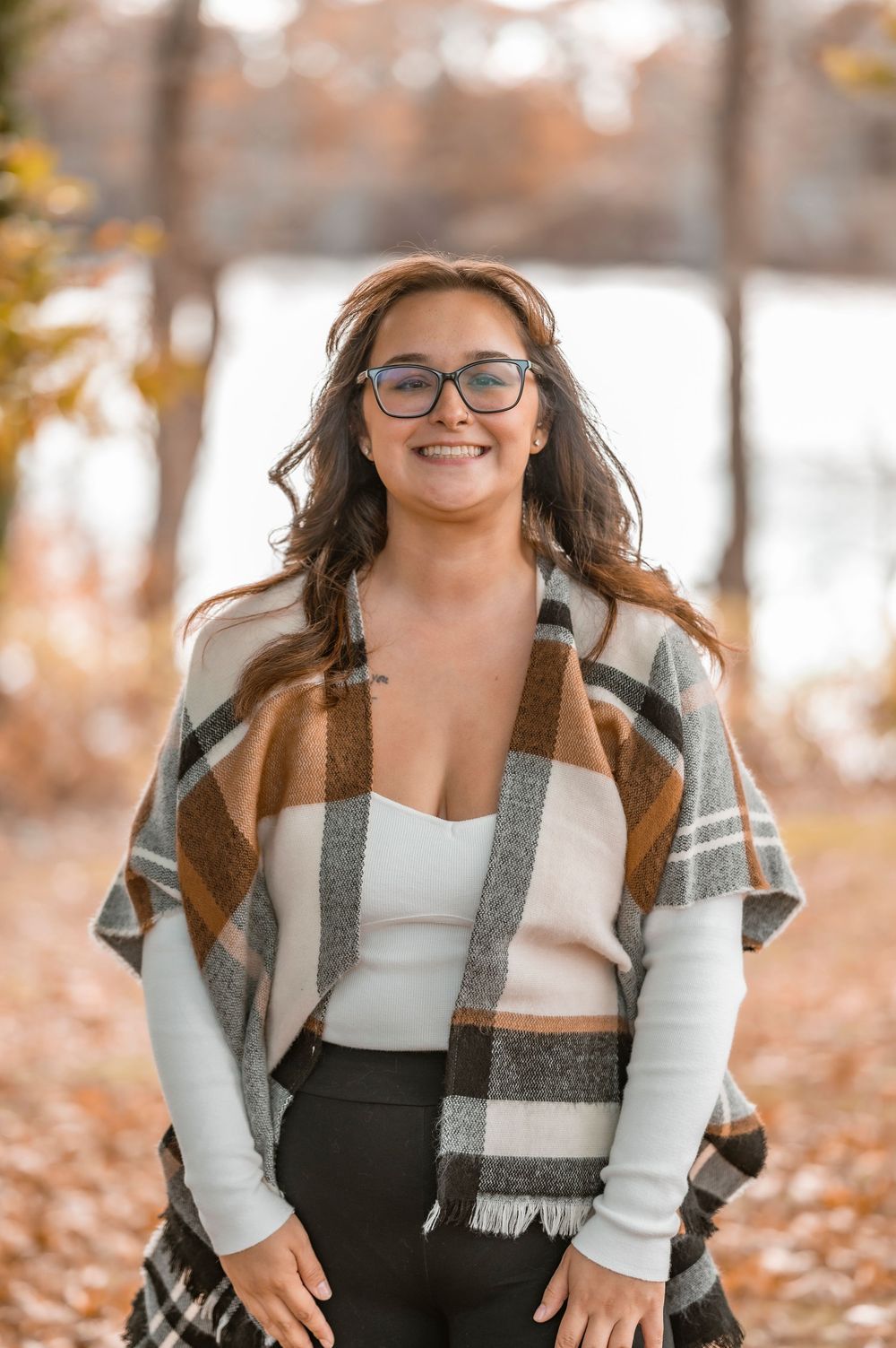A person wearing glasses and a plaid shawl smiles warmly while standing among fallen autumn leaves in a park setting.