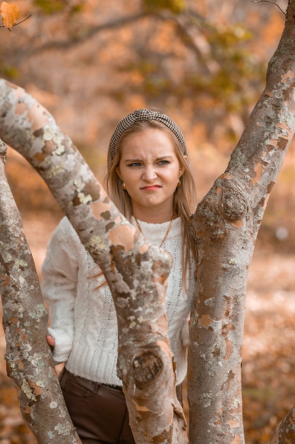 A person in a white sweater and headband poses between two tree branches on an autumn day.