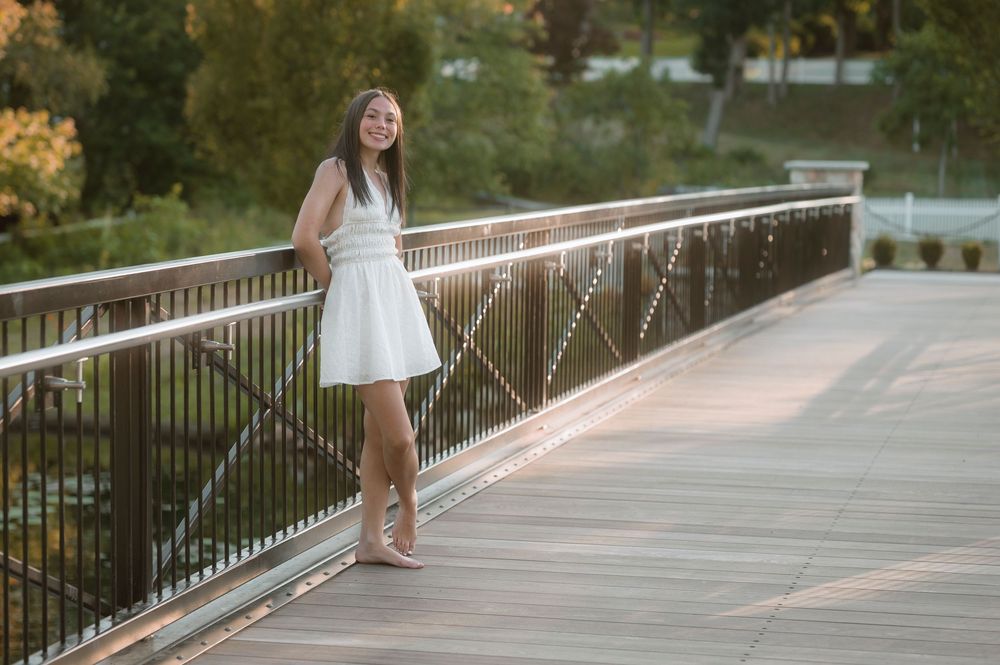 A young woman in a white dress stands confidently on a bridge in an outdoor summer setting.
