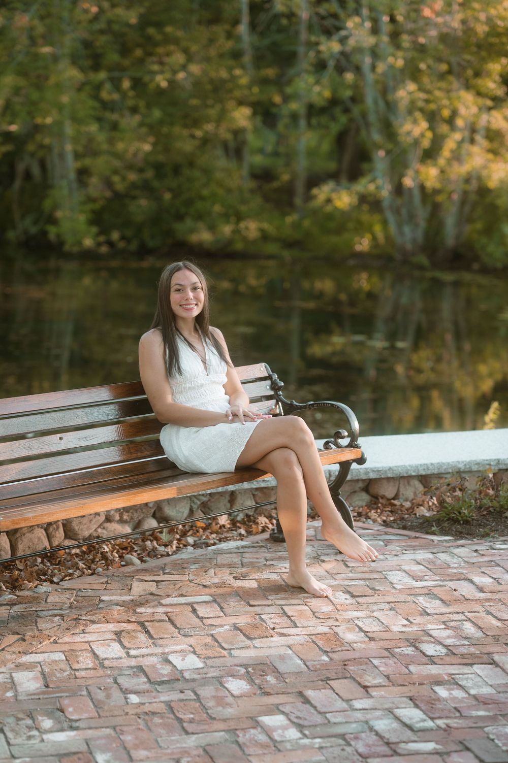 A white dress adorns a figure seated on a brick patio bench by a tranquil pond in autumn.
