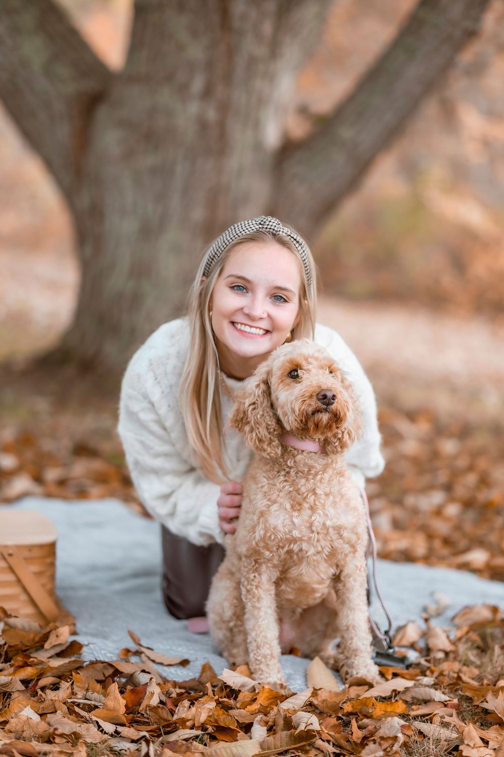 A goldendoodle dog sits on a blanket among autumn leaves with a companion wearing a cream sweater.