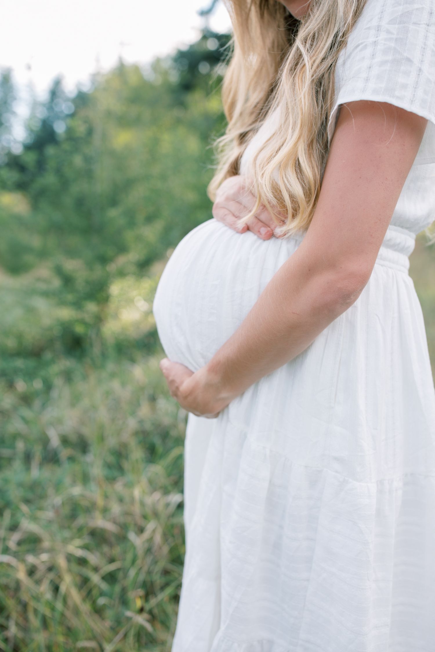 Pregnant woman in a white dress cradling her baby bump in a green field.