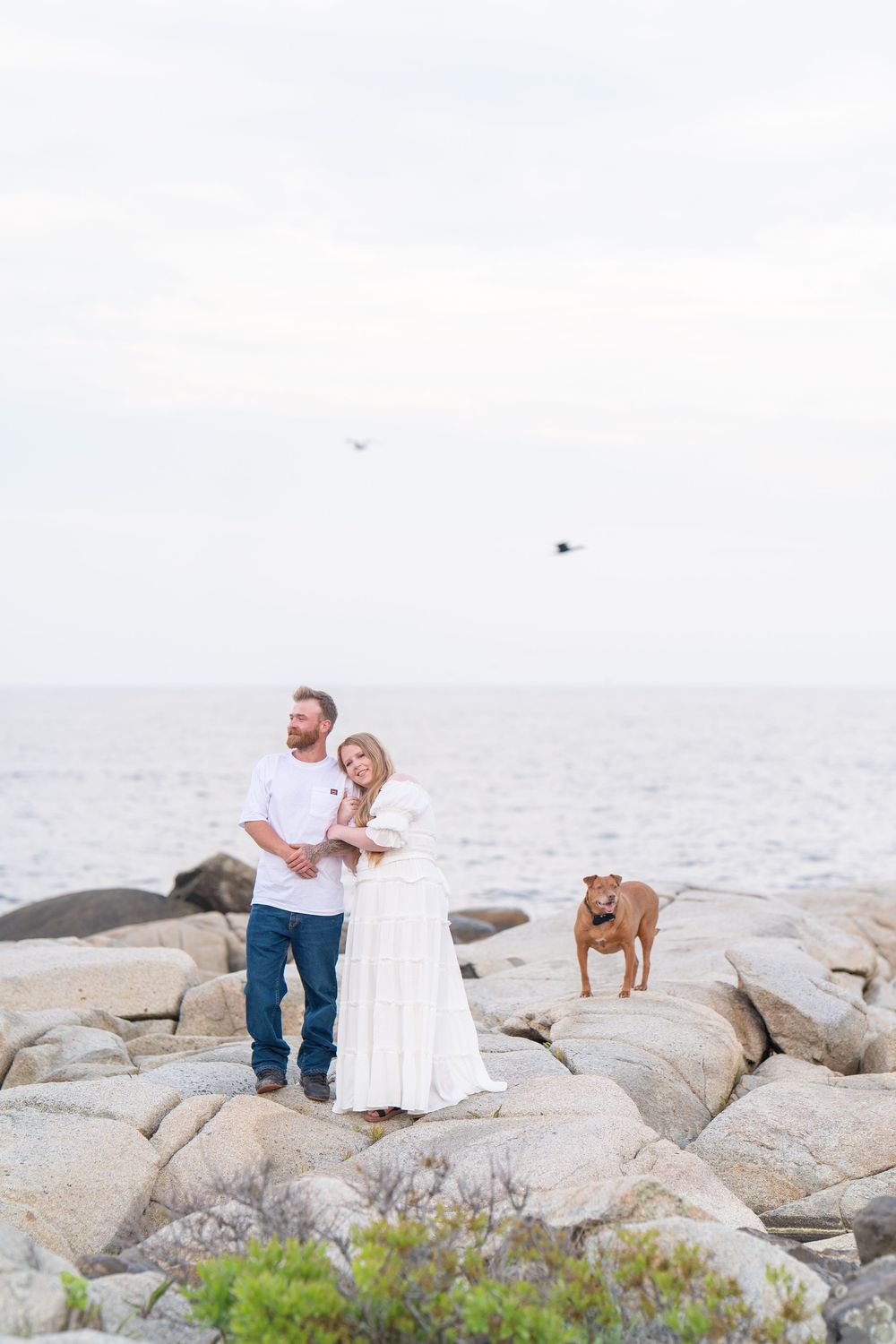 A couple in white and jeans stands on rocky shoreline with their brown dog near the ocean on a cloudy day.