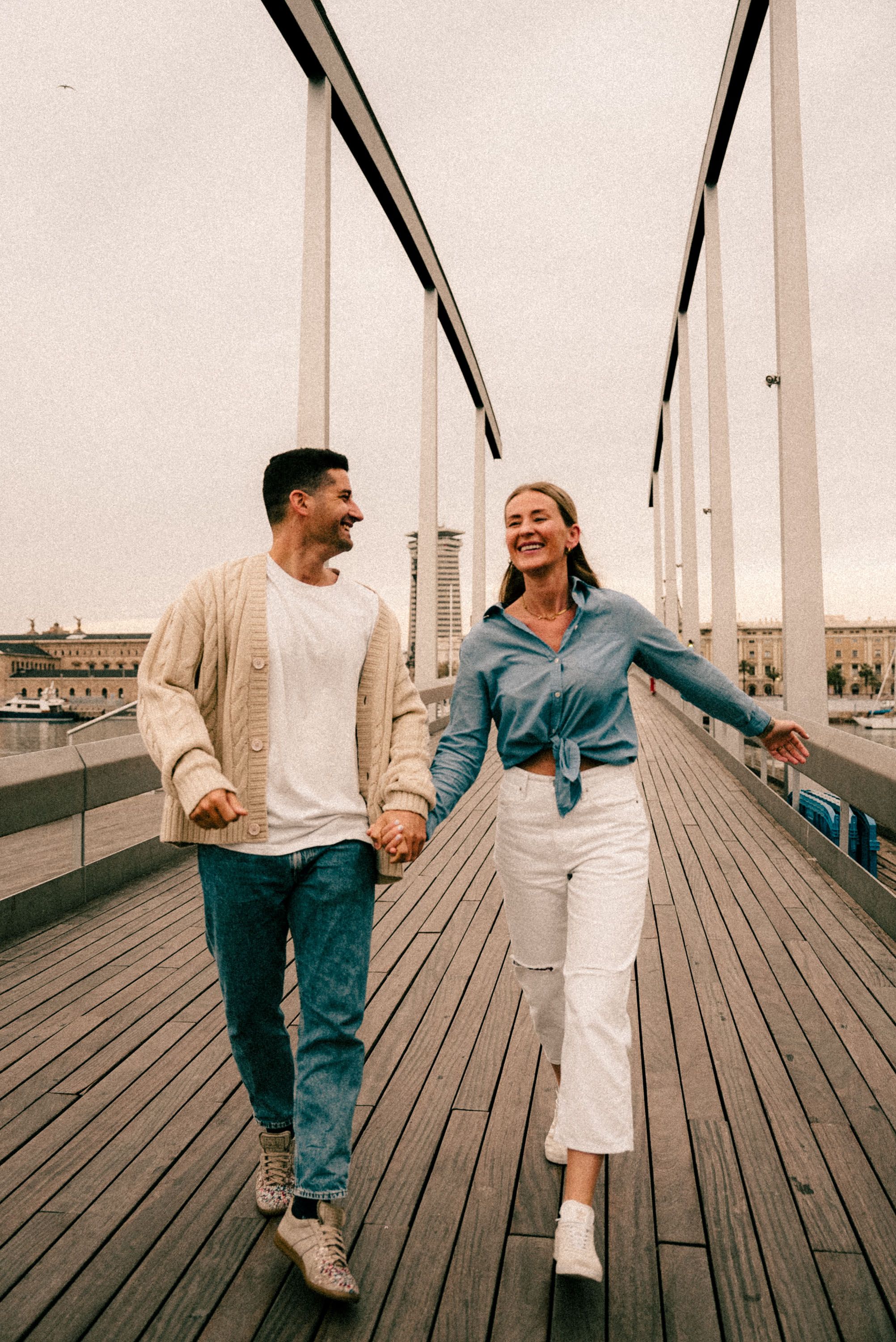 A couple walks hand in hand on a wooden boardwalk pier, wearing casual outfits and smiling.