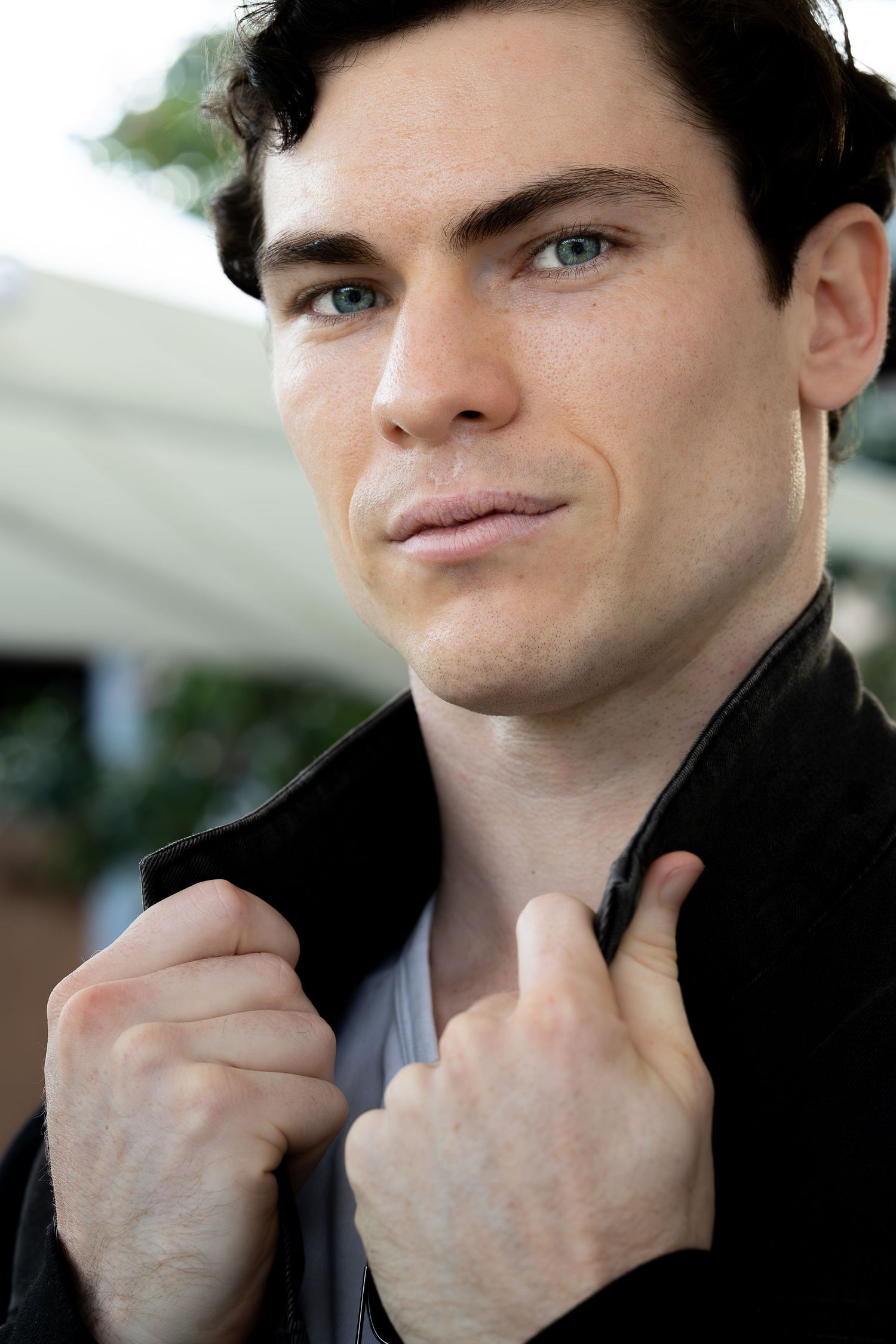 Handsome young man with blue eyes holding black jacket collar, looking confidently at camera outdoors.