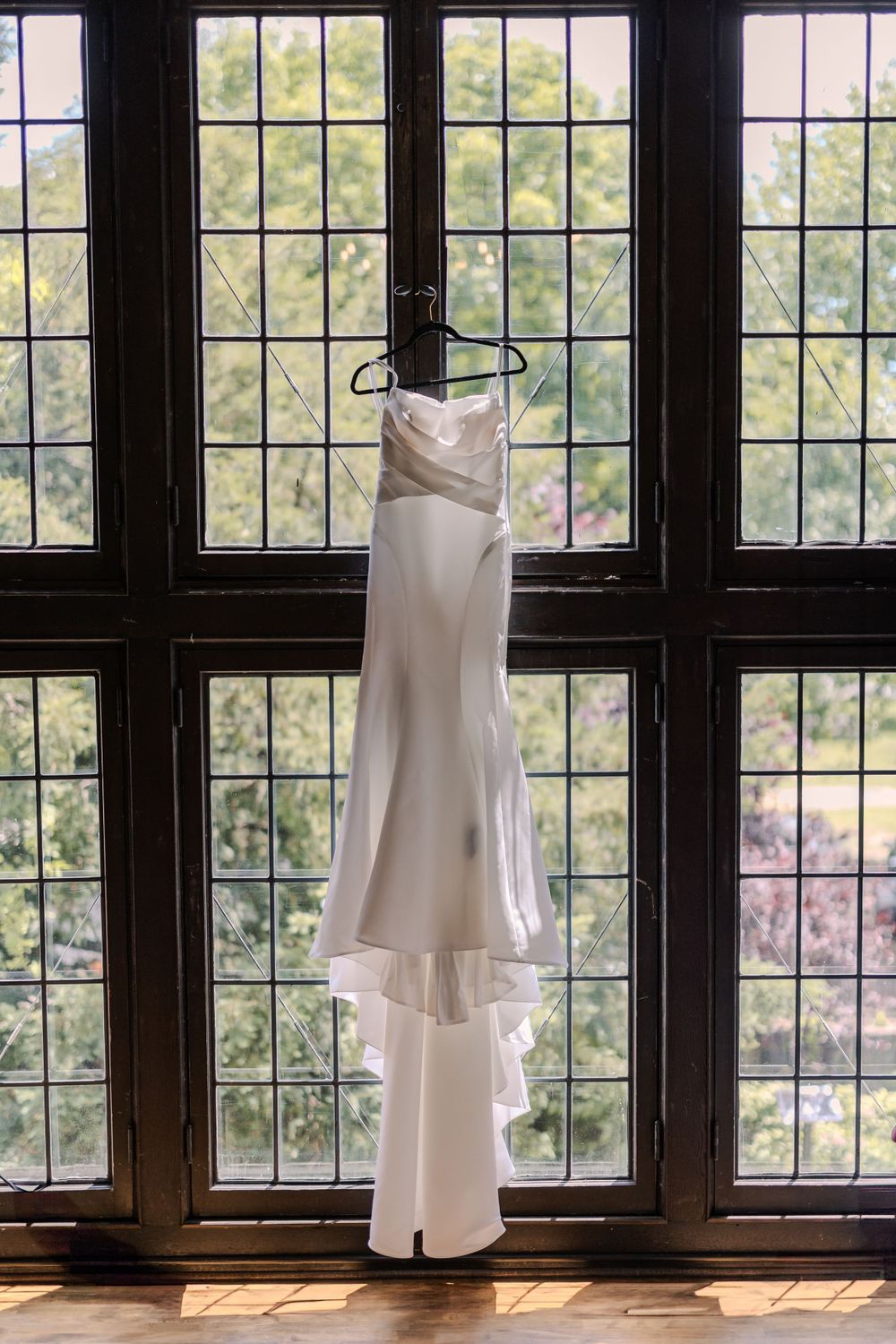 White wedding dress hanging in front of tall window with natural lighting.