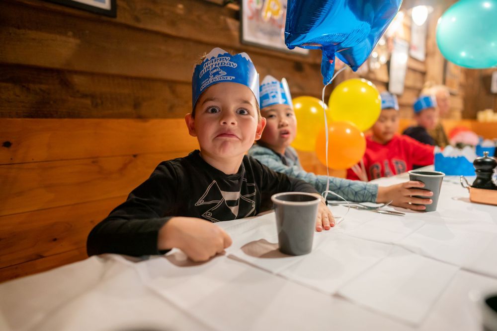 Joyful 7th birthday party photography of a boy blowing out candles with friends in a Snowplanet party room, by an Auckland photographer.