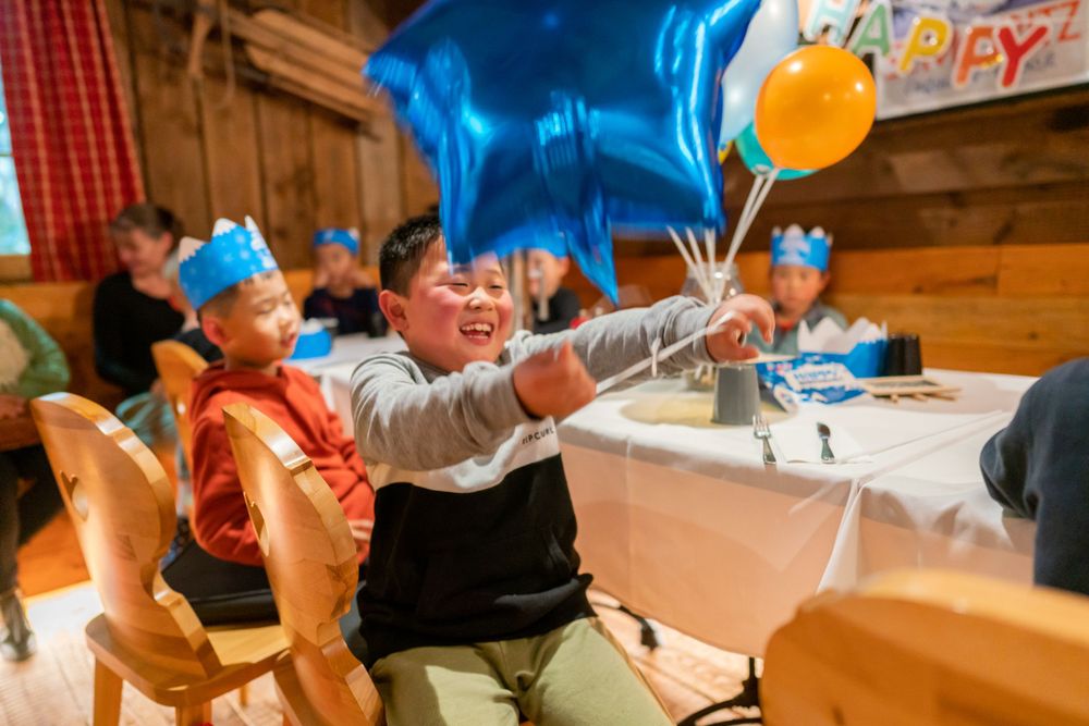 Joyful 7th birthday party photography of a boy blowing out candles with friends in a Snowplanet party room, by an Auckland photographer.