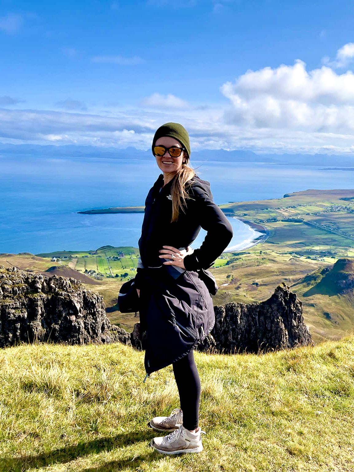Hiker stands on scenic coastal cliffs overlooking blue ocean and rolling green hills on a sunny day.