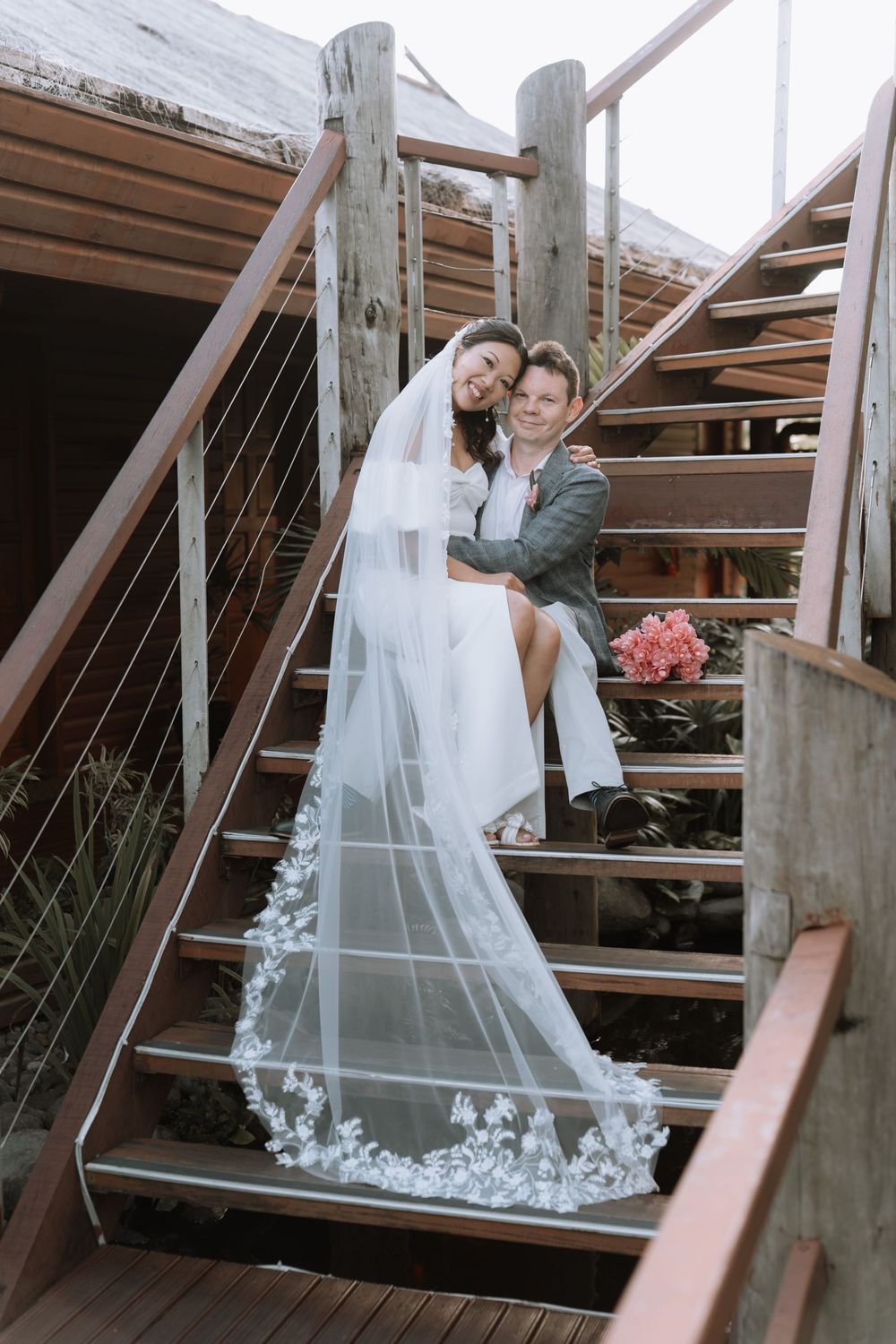 A newlywed couple sits together on wooden stairs, with a long flowing embroidered veil cascading down the steps.