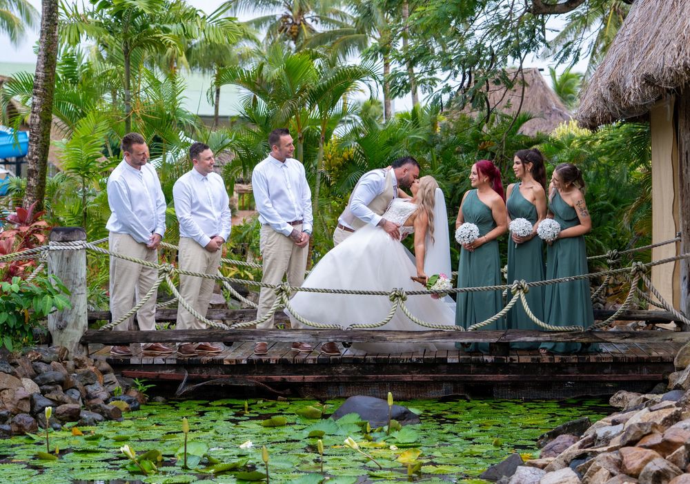 Wedding party stands on wooden bridge over lily pond at tropical resort with thatched roof huts and palm trees.